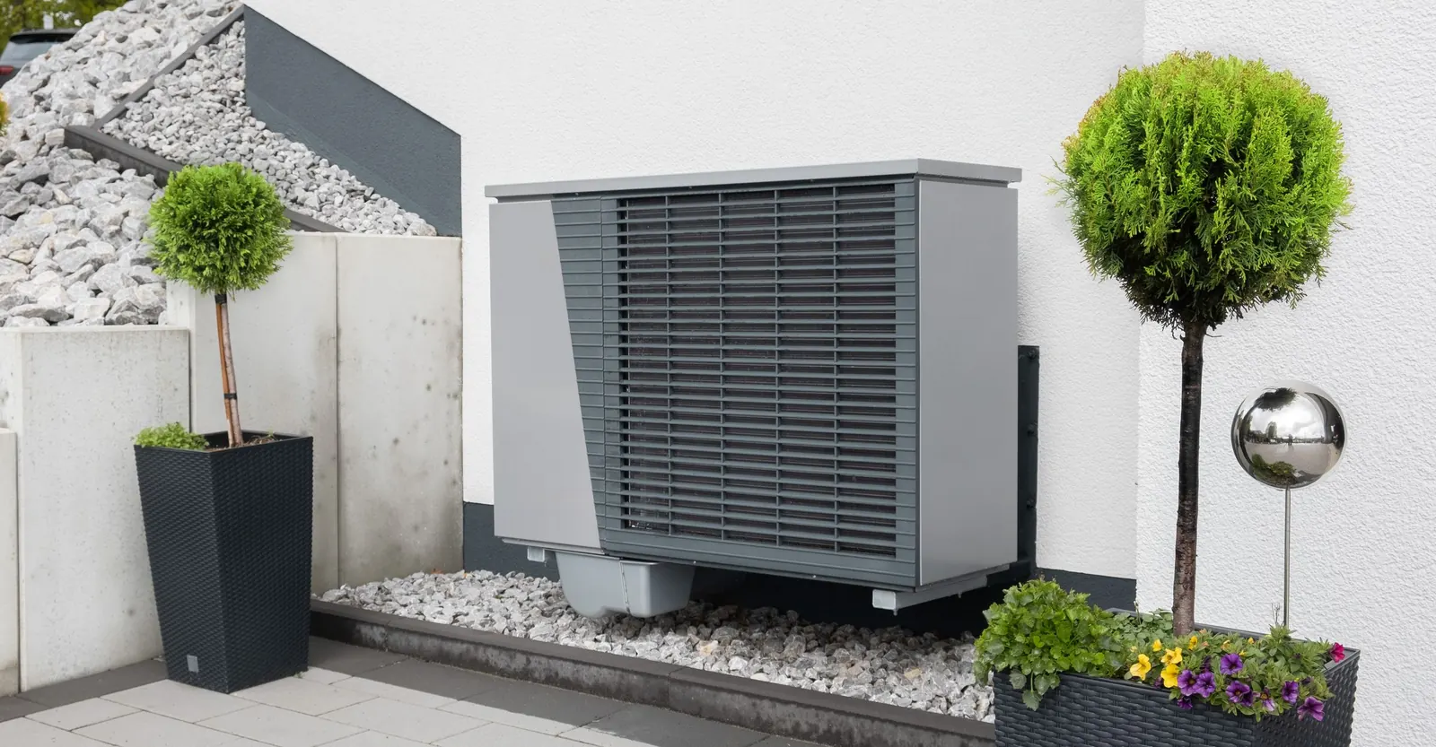 Pompa di calore grigia contro un muro esterno bianco, su un letto di sassi chiari